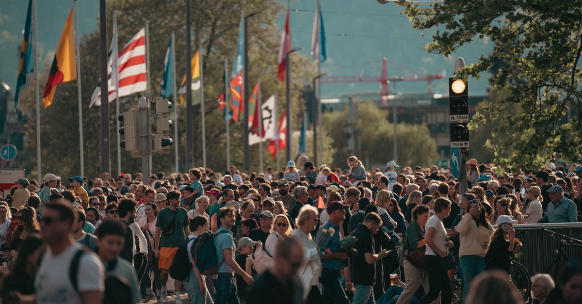 A bustling city street with a diverse crowd and international flags under a sunny sky.